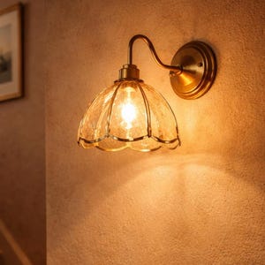 May include: A wall-mounted light fixture with a clear, textured glass shade and a brass-colored metal frame. The light is on, casting a warm glow on the textured wall. The fixture is mounted on a wall next to a staircase.
