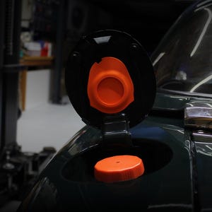 May include: A dark green vehicle fuel filler door is open, revealing a bright orange fuel cap. The interior of the circular fuel door lid also features a matching orange circular insert. The car body is glossy and reflects the surrounding garage light.
