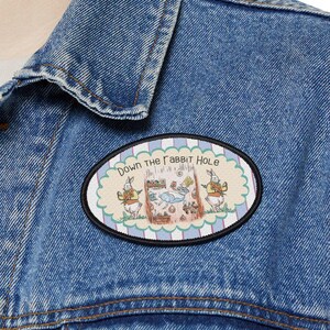 May include: A blue denim jacket with an oval patch. The patch displays the text "Down the Rabbit Hole" and a colourful illustration of rabbits and other elements, set within a light blue border.