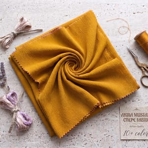 May include: Mustard yellow crepe fabric, artfully arranged, with a textured surface. The image includes a spool of thread, decorative scissors, and a tag that reads "Akira Mustard Crepe Fabric, 10+ colors."
