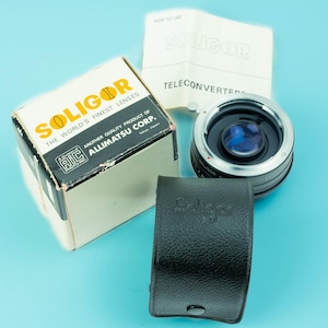May include: A Soligor teleconverter kit, including a lens, box, case, and instructions. The box is cream-colored with the Soligor logo and text. The lens is black and silver, and the case is black leather. The instructions are white with black text.