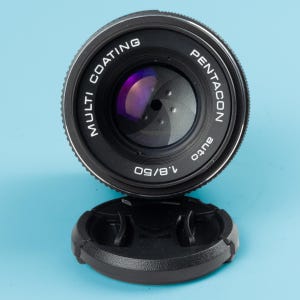 May include: A black Pentacon Auto 1.8/50 camera lens with a lens cap. The lens has the words "Multi Coating" and "Pentacon" printed on it. The lens cap is black and sits below the lens on a blue background.