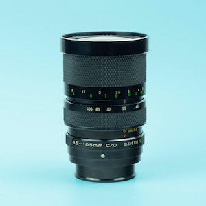 May include: A black 35-105mm C/D camera lens with a textured grip. The lens has various markings, including focal lengths and aperture settings. The lens is cylindrical with a macro setting and a series of numbers.
