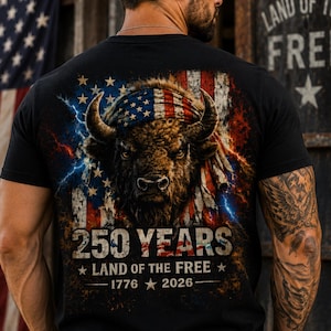 Può includere: Una t-shirt nera con la grafica di un bisonte che indossa una bandana con la bandiera americana. Lo sfondo mostra una bandiera americana invecchiata con effetti di fulmini. Il testo recita: 250 YEARS, LAND OF THE FREE, 1776, 2026, con stelle.