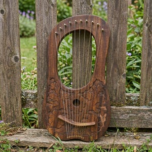 May include: A handcrafted wooden lyre with a dark brown finish, featuring intricate floral carvings. The instrument has 10 strings and a curved soundboard. It is leaning against a weathered wooden fence.