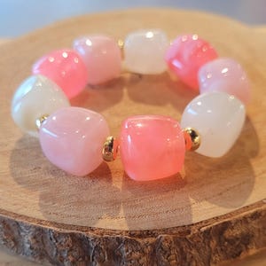 Peut inclure: Un bracelet composé de perles arrondies et translucides roses et blanches. De petits éléments métalliques dorés séparent les perles. Le bracelet est présenté sur une surface en bois. Les perles mesurent environ 1 cm de diamètre.