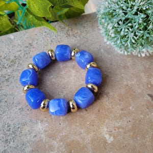 Peut inclure: Un bracelet de perles bleues avec des accents dorés. Le bracelet est composé de perles carrées bleues translucides séparées par de petites perles dorées. Le bracelet est présenté sur une surface texturée, avec de la verdure en arrière-plan.