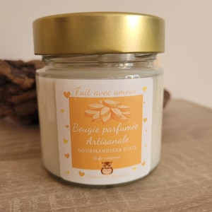 Puede incluir: Una pequeña vela perfumada artesanal en tarro de cristal con tapa dorada. La vela tiene cera blanca y una etiqueta naranja y blanca con un diseño floral y un búho. El texto incluye "Fait avec amour" y "Bougie parfumée Artisanale".