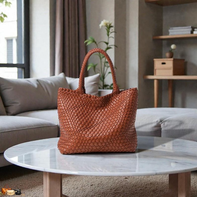 May include: A woven brown leather tote bag with two handles sits on a white marble coffee table. The bag has a rectangular shape and a textured surface. The background includes a sofa and a plant.