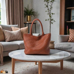 May include: A woven brown leather tote bag with a structured design. The bag has a large main compartment and two top handles. The bag is sitting on a round marble coffee table.