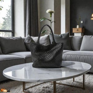 May include: A black woven tote bag with two handles sits on a marble-topped coffee table. The bag has a basket-weave design and is in front of a gray sectional sofa. The table has a gold-colored metal base.