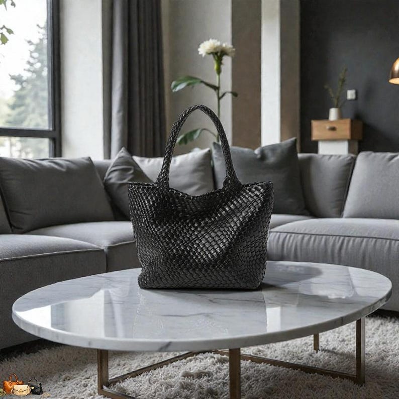 May include: A black woven tote bag with two handles sits on a marble-topped coffee table. The bag has a basket-weave design and is in front of a gray sectional sofa. The table has a gold-colored metal base.