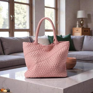 May include: A light pink woven tote bag with braided handles. The bag is rectangular with a wide base and a woven pattern. The bag is sitting on a white marble table.