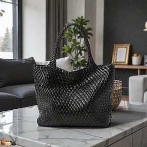 May include: A black woven tote bag with two handles. The bag is made of a tightly woven material, creating a textured surface. The bag is sitting on a marble table in a well-lit room. The bag is a stylish accessory.
