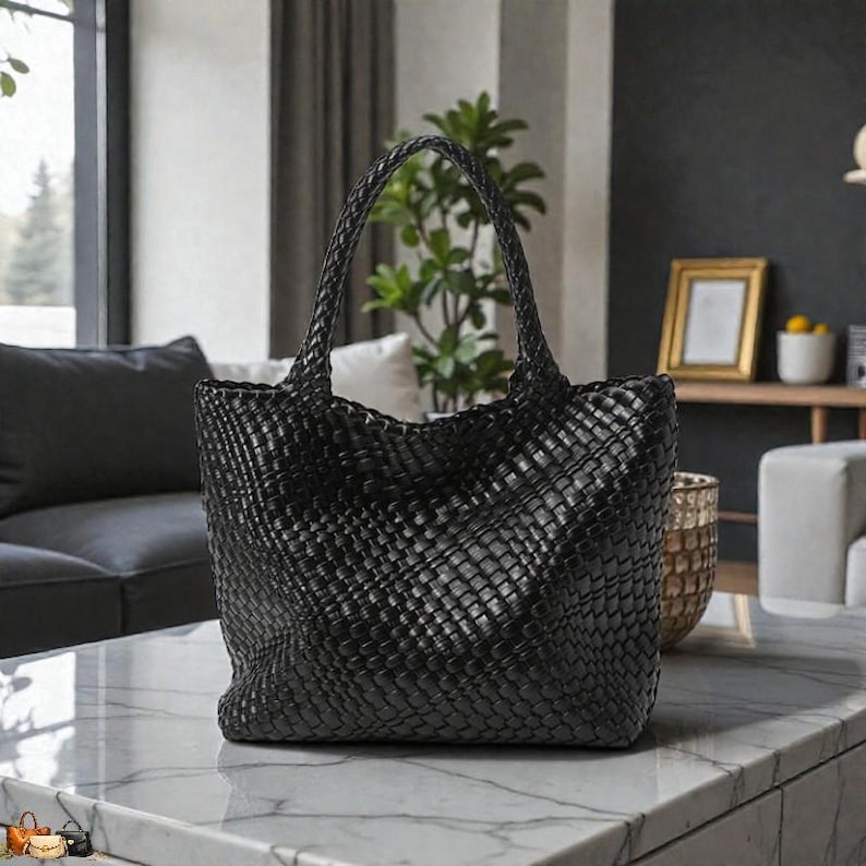May include: A black woven tote bag with two handles. The bag is made of a tightly woven material, creating a textured surface. The bag is sitting on a marble table in a well-lit room. The bag is a stylish accessory.