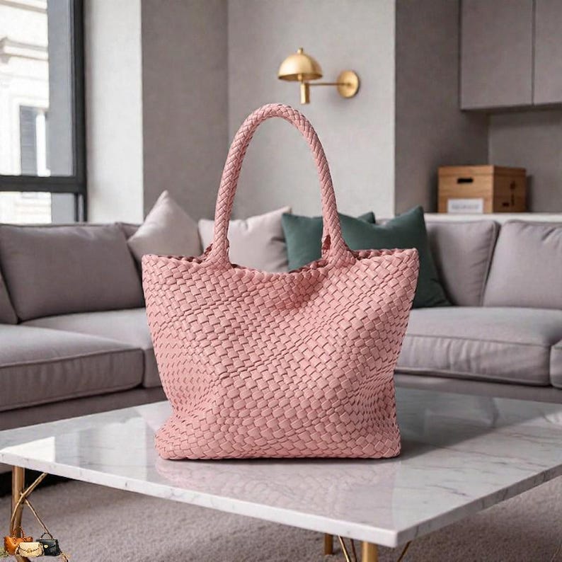May include: A woven pink tote bag with a braided handle sits on a marble coffee table. The bag has a rectangular shape and a textured surface. The background includes a sofa with pillows and a wall-mounted light fixture.