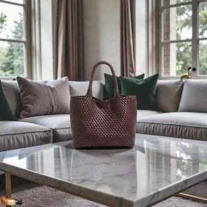 May include: A woven tote bag in a deep burgundy hue sits on a marble coffee table. The bag features a structured shape with a woven pattern and a top handle. The background includes a sofa with pillows and a window.