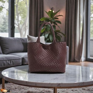 May include: A woven tote bag in a deep burgundy color. The bag features a structured design with a rectangular base and a woven pattern throughout. It has two sturdy handles for carrying. The bag is sitting on a marble table.