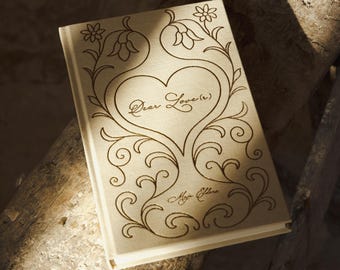 May include: A beige journal with a decorative floral and heart design. The cover features the words "Dear Love" and the artist's signature. The journal is resting on a weathered wooden surface.