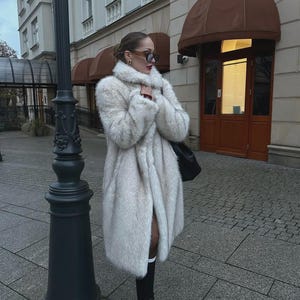 May include: A woman wearing a long, off-white faux fur coat, black boots, and sunglasses stands on a paved street. The coat is open, revealing a glimpse of a red top. A black bag hangs over her shoulder. The background includes a building and street lamp.