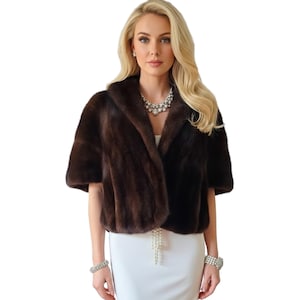 May include: A brown fur bolero with short sleeves. The bolero has a shawl collar and a luxurious appearance. The garment is worn over a white dress. The model is wearing a necklace and bracelets.