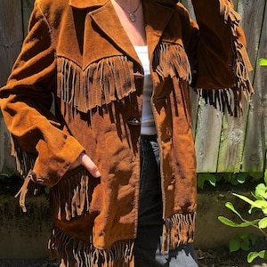 Puede incluir: Una chaqueta de ante marrón con flecos largos en el pecho, las mangas y el dobladillo. La chaqueta tiene cuello, mangas largas y se abrocha con tres botones de palanca de color marrón oscuro en la parte delantera.