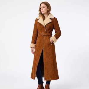 May include: A long, tan suede coat featuring a cream-colored faux shearling lapel and cuffs. The coat has a full-length front zipper, a matching belt with a rectangular buckle at the waist, and two side pockets. It is worn over dark blue jeans.
