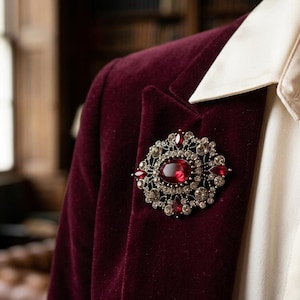 Könnte beinhalten: Nahaufnahme eines bordeauxfarbenen Samtblazers mit einem weißen Hemd mit Kragen. Eine große, verzierte Brosche mit einem zentralen roten Stein und klaren Kristallen ist an dem Blazer befestigt.