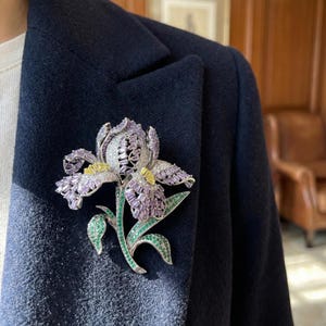 Peut inclure: Une broche détaillée en forme de fleur d'iris, ornée de pierres précieuses violettes, blanches, jaunes et vertes. La broche est fixée à un manteau bleu marine foncé. Les pétales de la fleur sont un mélange de pierres violettes et blanches.