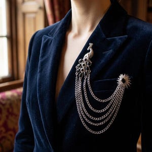 Peut inclure: Un blazer en velours bleu marine orné de deux broches argentées. L'une est un paon avec des détails noirs et des chaînes pendantes. L'autre est un motif de soleil avec un centre noir. Le blazer est porté par une personne.