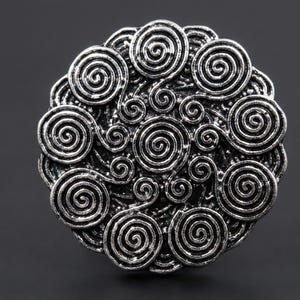 Peut inclure: Un broche circulaire argenté avec un motif en spirale détaillé. La broche présente de nombreuses spirales concentriques de tailles variables, créant un aspect texturé et ornemental. Le métal a une finition vieillie et antique.