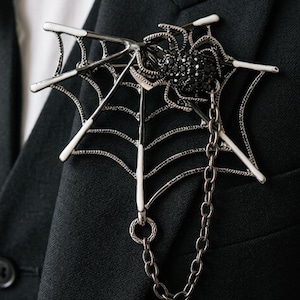 以下が含まれることがあります： 黒いクモと鎖が付いた、銀色のクモの巣のブローチ。クモの体は黒い宝石でできています。ブローチは黒いブレザーに取り付けられており、ゴシックまたはハロウィーンをテーマにしたアクセサリーです。