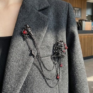 Peut inclure: Ensemble de broches gris foncé comprenant un oiseau perché sur une pierre précieuse rouge et une cage à oiseaux avec des roses rouges. La broche a des chaînes pendantes et des gemmes rouges en forme de goutte. La broche est fixée à un blazer gris.