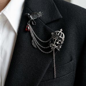 Può includere: Spilla argentata con un corvo, una gemma rossa e un design a corona. La spilla è attaccata a un blazer nero con catene e dettagli di piume, creando un'estetica gotica.