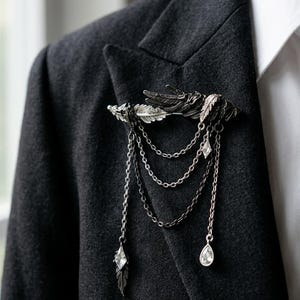 Peut inclure: Broche argentée ornée d'une plume et de chaînes, ornée de cristaux transparents. La broche est fixée à un blazer gris foncé, mettant en valeur ses détails complexes et son style élégant. Les chaînes pendent gracieusement.