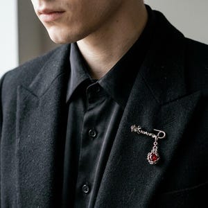 Pode incluir: Um broche prateado com uma pedra vermelha pendurada é preso a um blazer preto. O broche tem um design detalhado e ornamentado. A pessoa está usando uma camisa preta abotoada por baixo do blazer. A estética geral é escura e elegante.