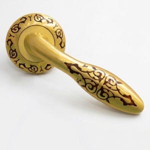 Pode incluir: Uma maçaneta dourada com intrincados desenhos florais castanho-escuros. A maçaneta apresenta uma alavanca curva e uma placa traseira circular, mostrando padrões detalhados. A ferragem está sobre um fundo branco.
