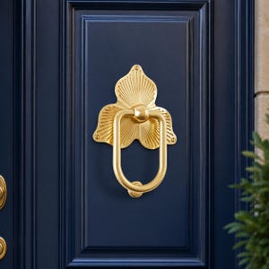 Può includere: Un battente per porta in metallo color oro lucido caratterizzato da una piastra decorativa a forma di ventaglio con scanalature lineari radianti e un anello oscillante liscio a forma di U. La ferramenta è montata su una porta blu scuro.