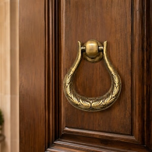 May include: A teardrop-shaped brass door knocker with an intricate embossed leaf pattern mounted on a dark brown wooden door. The metal hardware features a polished antique finish with a hinged ring mechanism attached to a circular backplate.