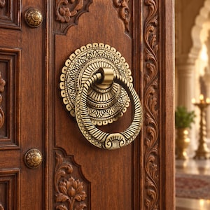 May include: A brass door knocker with a circular ring and an ornate, scalloped backplate featuring intricate floral engravings. The knocker is mounted on a dark brown wooden door with carved panels and two small, matching brass decorative studs.