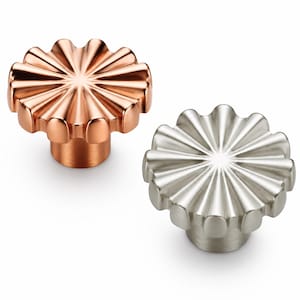 May include: Two circular cabinet knobs with a fluted, sunburst-like design radiating from the center. One knob features a polished copper finish, while the other has a brushed silver-toned metallic finish, both with a cylindrical base.