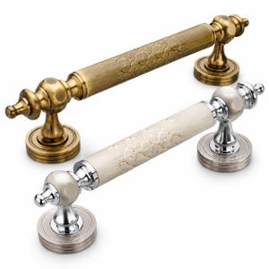 May include: Two cylindrical door handles with decorative circular bases. One handle features an antique brass finish with an engraved floral pattern. The second handle has a white ceramic center with a gold floral pattern and polished chrome ends.
