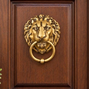 May include: A brass-colored metal door knocker shaped like a lion head with a circular ring held in its mouth. The lion features detailed facial features and a flowing mane, mounted on a dark brown wooden door panel with a rectangular frame.