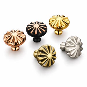 Könnte beinhalten: Eine Sammlung von fünf runden Schrankknöpfen mit einem Strahlenmuster und erhabenen Rippen. Das Set enthält Ausführungen in poliertem Kupfer, Mattschwarz mit Kupferakzenten, poliertem Gold, gebürstetem Messing und gebürstetem Silber.