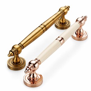May include: Two door handles with cylindrical grips and circular mounting plates. One handle features an antique brass finish, while the other combines a polished rose gold metal finish with a smooth, cream-colored central cylindrical insert.