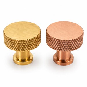 Puede incluir: Dos pomos cilíndricos para armarios con parte superior plana y borde moleteado con patrón de diamante. Un pomo tiene un acabado en oro cepillado, mientras que el otro presenta un acabado en cobre cepillado. Ambos tienen una base circular lisa.