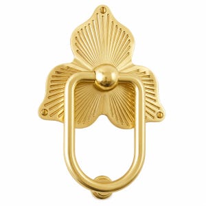 May include: A polished gold-tone metal door knocker featuring a decorative backplate shaped like a stylized flower with radiating sunburst lines. The assembly includes a smooth, elongated U-shaped ring and two visible mounting screws at the top and base.