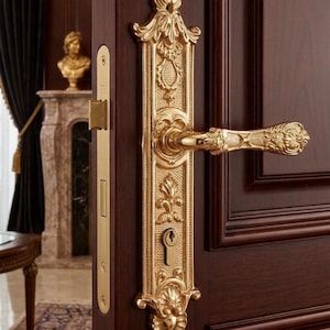 May include: A detailed gold-coloured door handle and lock set, mounted on a dark brown wooden door. The set features intricate floral and scrollwork patterns. The handle and lock have a vintage, antique appearance, with a keyhole visible.