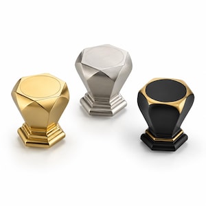 May include: Three hexagonal cabinet knobs with a faceted geometric design. One knob is polished gold, one is brushed silver, and one is matte black with gold accents. Each knob features a flat circular top and a tiered hexagonal base.