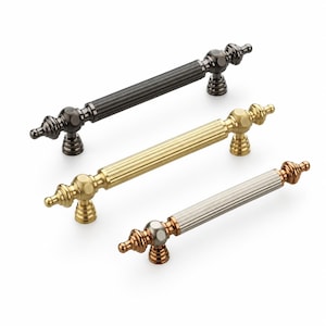 May include: Three cabinet pulls arranged diagonally, each featuring a central fluted cylindrical bar and decorative finials at both ends. The top pull is dark gunmetal, the middle is polished brass, and the bottom is a mix of copper and silver.